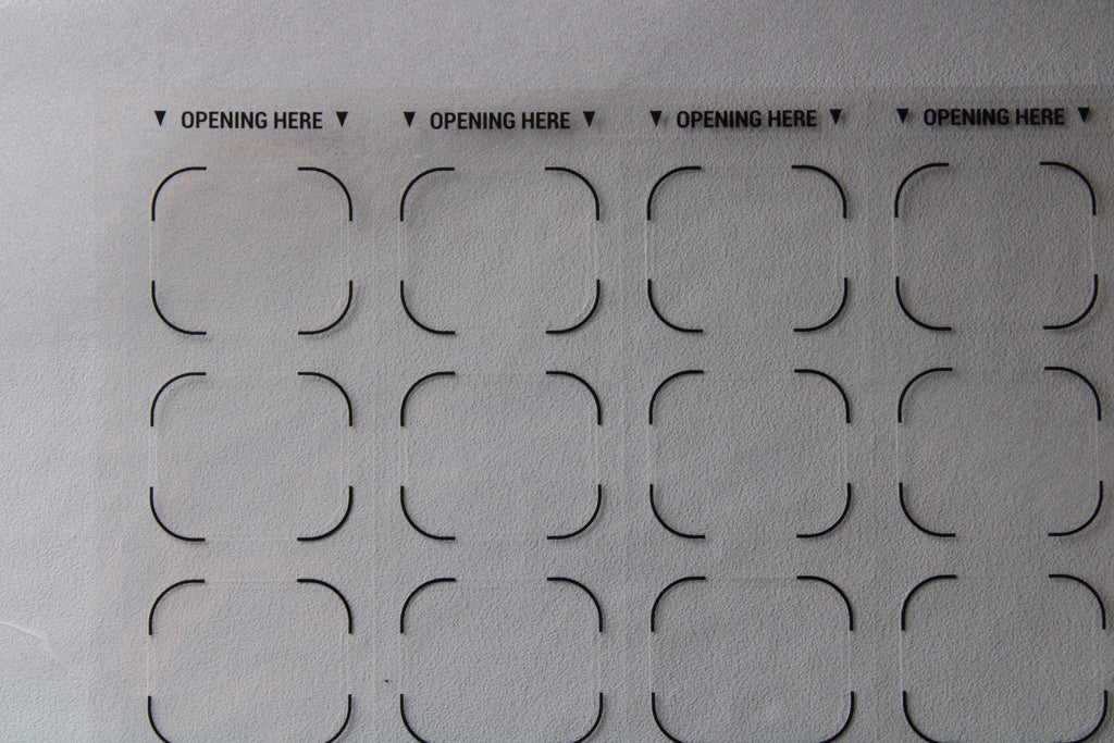 Sheet of pre-cut cardboard shapes with 'OPENING HERE' text on a gray background. Correction labels for pharmacy blister packaging and medication labeling