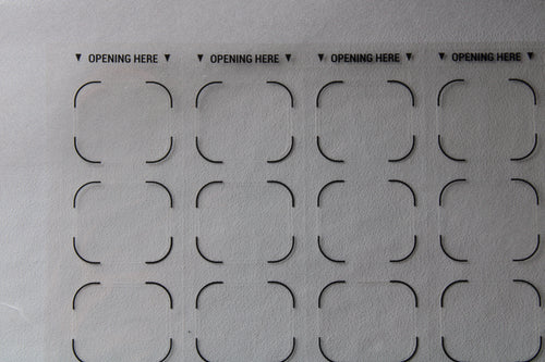 Sheet of pre-cut cardboard shapes with 'OPENING HERE' text on a gray background. Correction labels for pharmacy blister packaging and medication labeling